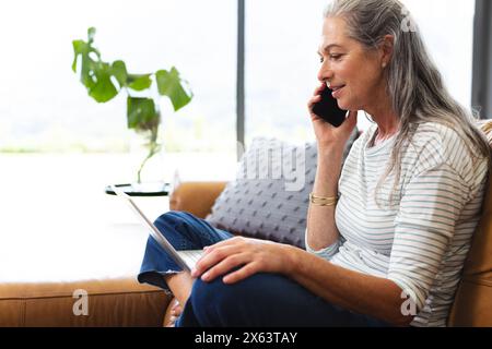 À la maison, femme caucasienne mature parlant au téléphone, tenant une tablette Banque D'Images