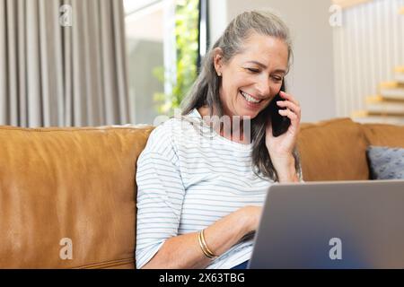 À la maison, femme mature caucasienne parlant au téléphone, assise près d'un ordinateur portable Banque D'Images