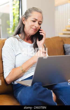 À la maison, femme caucasienne mature parlant au téléphone, en utilisant un ordinateur portable Banque D'Images