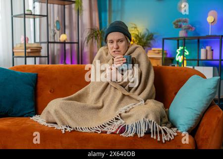 Femme blonde couverte par une couverture chaude boit du café à la maison. La femelle caucasienne gèle à cause du froid, se sent mal, malsaine. fille malade avec des symptômes de la grippe couverte de couverture se réchauffe avec du thé chaud à la maison Banque D'Images