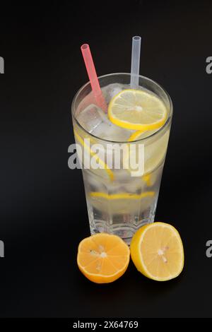 Limonade rafraîchissante dans un grand verre à facettes avec de la glace, des pailles et des tranches de citron sur un fond noir. Gros plan. Banque D'Images