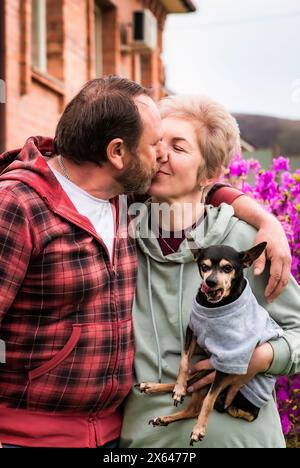 Couple marié russe mature embrassant et serrant près du buisson fleuri de rhododendron, tenant un chien Banque D'Images