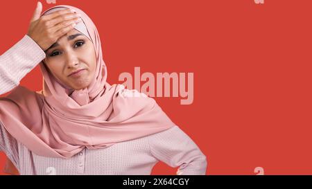 Oups oublie. Regret d'erreur. Femme bouleversée frustrée déçue en foulard hijab touchant la tête avec la main isolée sur fond d'espace vide rouge. Banque D'Images