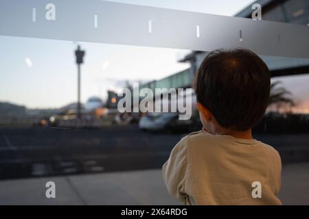 Un jeune enfant regarde par une fenêtre dans un aéroport très fréquenté. La scène est animée par l'activité, avec des avions décollant et atterrissant, des voitures roulant sur le Banque D'Images