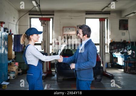 Mécanicien automobile féminin parlant avec le client, serrant la main. Belle femme travaillant dans un garage, portant des combinaisons bleues. Technicien d'entretien automobile féminin Banque D'Images
