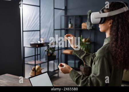 Jeune femme avec de longs cheveux bouclés debout dans le salon en utilisant un casque VR, espace de copie Banque D'Images