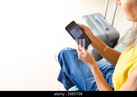 À la maison, femme caucasienne mature tenant un smartphone, assise à côté de la valise, espace de copie Banque D'Images