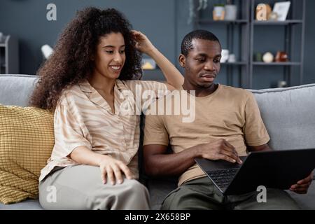 Jeune couple moderne ethniquement diversifié assis sur le canapé dans le salon à la maison, surfer sur Internet sur ordinateur portable Banque D'Images