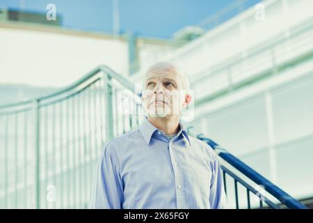 Homme mature se tient en toute confiance à l'extérieur, chemise bleue améliorant son comportement sage dans un contexte architectural moderne, exsudant le professionnalisme Banque D'Images