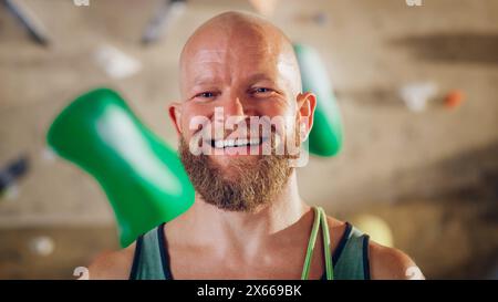 Athlète masculin masculin fort souriant et posant au Rock Climbing Gym avec fond de mur de Bouldering. Beau homme chauve heureux avec Portrait de barbe de gingembre, portant un maillot de corps coloré. Banque D'Images