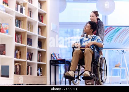 Portrait en longueur d'un adolescent handicapé choisissant des livres dans la bibliothèque de l'école avec une fille souriante aidant l'espace de copie Banque D'Images