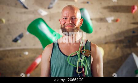 Athlète masculin masculin fort souriant et posant au Rock Climbing Gym avec fond de mur de Bouldering. Beau homme chauve heureux avec barbe de gingembre, portant un maillot de corps coloré et tenant un harnais de corde. Banque D'Images