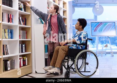 Portrait de vue de côté de deux adolescents dans la bibliothèque de l'école avec garçon en fauteuil roulant choisissant des livres sur l'espace de copie de l'étagère Banque D'Images