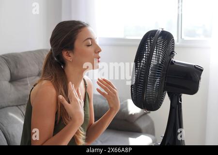 Chaleur estivale. Jeune femme hispanique se refroidissant par ventilateur à la maison, se sentant mal avec la température élevée par temps chaud. Fille latine devant Banque D'Images