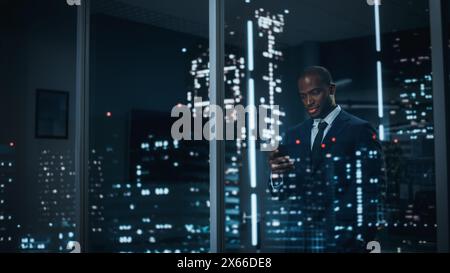 Homme d'affaires noir prospère dans un costume sur mesure debout dans son bureau regardant par la fenêtre sur Night City. Gestionnaire de placements performant travaillant tard. Prise de vue en extérieur élégante Banque D'Images
