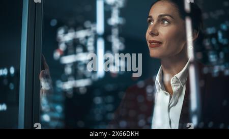 Bureau : femme d'affaires réussie en costume élégant travaillant, regardant dans l'émerveillement à Night City. PDG féminin élégant travaillant tard et dur pour créer une expérience d'achat en ligne e-commerce durable et sûre Banque D'Images