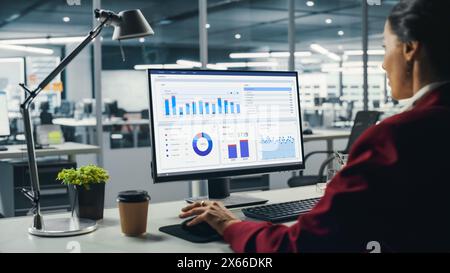 Femme d'affaires réussie assis au bureau travaillant sur ordinateur de bureau dans le bureau de la ville. Top Manager travaillant dur effectuant des recherches pour un projet de commerce électronique avec graphiques, graphiques et tableaux. Au-dessus de l'épaule Banque D'Images