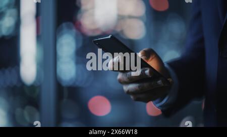 Bureau de nuit : homme d'affaires noir réussi portant costume debout, en utilisant smartphone. CEO navigation sur Internet, gestion de la stratégie des médias sociaux dans le logiciel de commerce électronique. Concentrez-vous sur la main avec le téléphone portable Banque D'Images