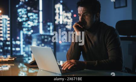 Portrait d'homme d'affaires stressé travaillant sur ordinateur portable dans son bureau de grande ville la nuit. Entrepreneur fatigué surchargé essayant de trouver une solution pour les problèmes d'affaires. Banque D'Images