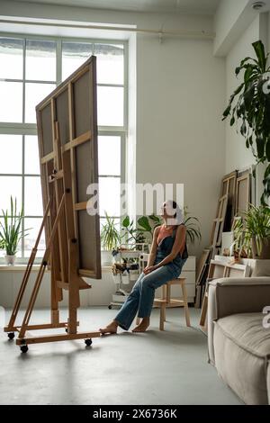 Artiste femme joyeuse regardant l'immense toile admirant l'examen assis à la maison studio d'art. Banque D'Images