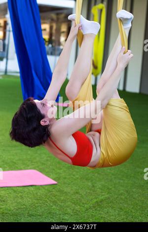 Une fille s'emmêle dans une posture drôle de tissu pendant le yoga aérien. Banque D'Images