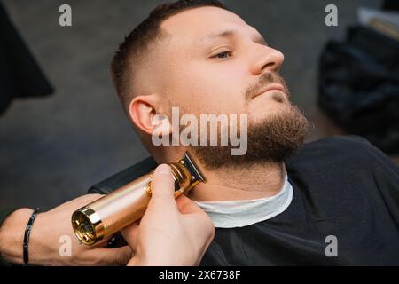 Styliste utilise une tondeuse et un peigne travaillant avec la barbe du client dans le gros plan de coiffeur. Barber coupe les cheveux sur le visage de l'homme avec la machine dans le salon de toilettage Banque D'Images