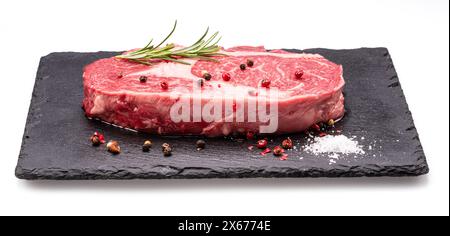 Steak de ribeye cru avec des grains de poivre et du romarin sur un plateau de service en graphite isolé sur fond blanc. Banque D'Images