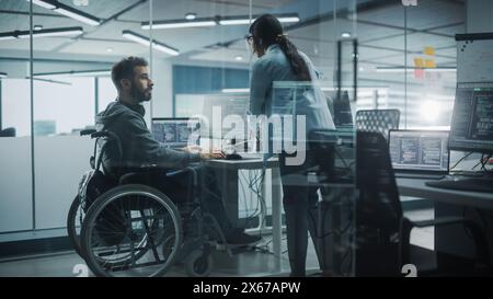 Bureau adapté aux personnes handicapées : Happy IT Programmer with Disability in a Wheelchair parle avec un collègue tout en travaillant à l'ordinateur. Les ingénieurs logiciels développent une application innovante, un programme. Banque D'Images