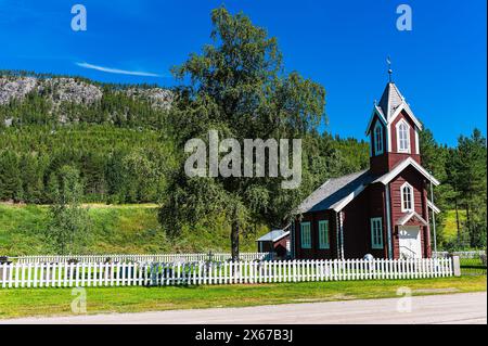 Une église en bois rouge est visible au premier plan avec une clôture blanche en arrière-plan. L'église se distingue par la verdure qui l'entoure. Banque D'Images