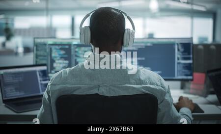 Bureau : Professional Black IT Programmer utilise un casque tout en travaillant sur un ordinateur de bureau. Développeur de site Web masculin, ingénieur logiciel développant une application, jeu vidéo. Écoute de podcast, musique. Vue arrière Banque D'Images