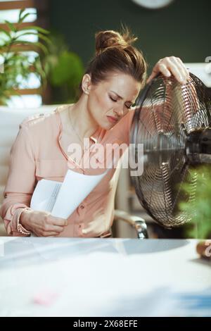 Lieu de travail durable. employée féminine moderne au travail avec des documents et un ventilateur électrique souffrant de la chaleur estivale. Banque D'Images