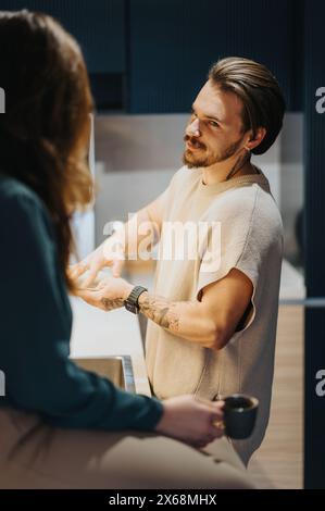 Jeunes collègues jouissant d'une atmosphère positive lors d'une pause matinale dans la cuisine du bureau, souriant et conversant tout en lavant la vaisselle ensemble. Banque D'Images