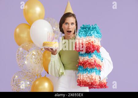 Jeune femme surprise dans le chapeau de fête avec gâteau d'anniversaire et pinata mexicaine sur fond de lilas Banque D'Images