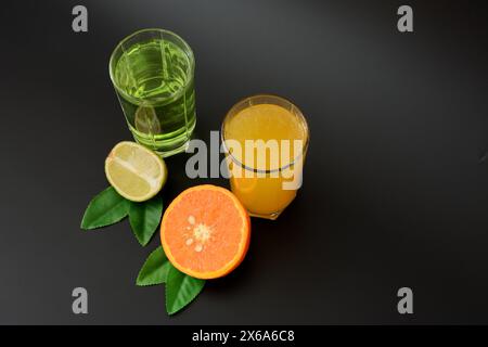 Deux grands verres avec différents jus d'agrumes sur un fond noir, à côté des moitiés d'orange mûr et de citron vert avec des feuilles. Gros plan. Banque D'Images