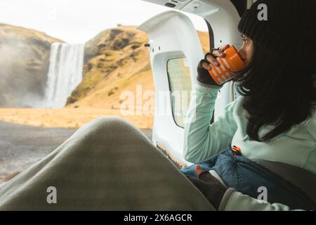 Femme touristique assis boire café profiter de la vue sur la nature avec porte ouverte à l'intérieur loué camping-car. Cascade de Skogafoss, Islande. Voyagez en solo à visiter l'islandais Banque D'Images