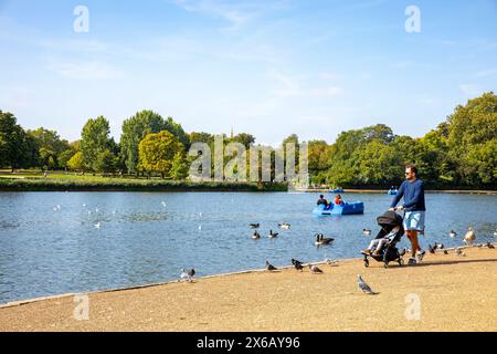 Hyde Park le Serpentine, jeune père pousse son enfant en poussette le long de la rive du lac, avec couple sur un pédalo, Londres, Angleterre, Royaume-Uni, 2023 Banque D'Images