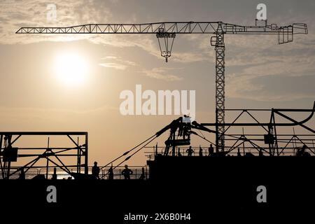 Sur le chantier de construction, il y a des ouvriers qui démantèlent la grue à tour et une autre grue à tour en attente de levage avec le coucher du soleil en mer. Banque D'Images