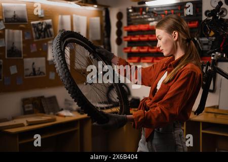 Jeune femme mécanicien de vélo fixant roue de cycle dans l'atelier de réparation. Ingénieur de magasin de vélo fixer la roue de vélo dans l'atelier. partie vélo dans les mains féminines. Banque D'Images