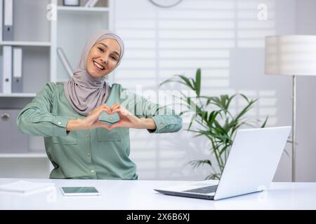 Une jeune femme musulmane joyeuse portant un hijab fait une forme de cœur avec ses mains, exprimant positivité et amour dans un cadre de bureau lumineux et contemporain. Banque D'Images