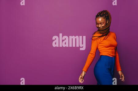 Fille heureuse avec des tresses vibrantes et bicolores se tient dans un studio. Elle porte des vêtements élégants et décontractés et un col polo tendance. L'arrière-plan violet ajoute Banque D'Images