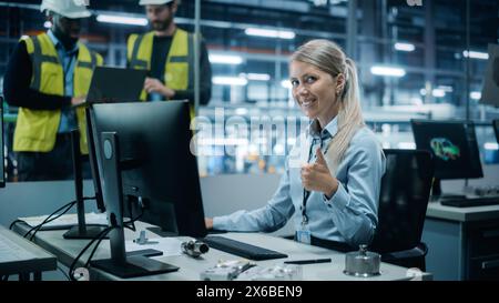 Bureau d'usine de voiture : Portrait d'une femme ingénieur travaillant sur un ordinateur de bureau, souriant et montrant le geste Thumbs Up. Équipe de techniciens en ligne de montage de bras robotisés automatisés fabrication électronique Banque D'Images