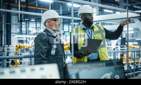 Usine de voiture : chef de projet et ingénieur automobile en casque dur, parler, utiliser un ordinateur portable. Surveillance, contrôle, production d'équipements. Chaîne d'assemblage de bras robotisés automatisée pour la fabrication de véhicules électriques Banque D'Images