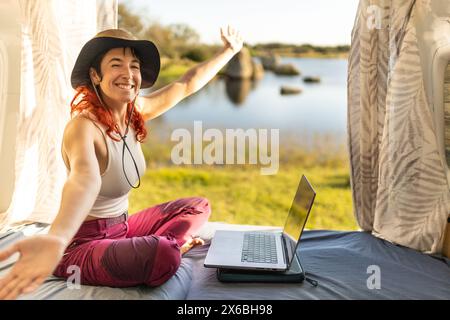 Femme aux cheveux rouges télétravail tapant avec ordinateur portable dans le camping-car au milieu de la nature près du lac, connecté à Internet, fille écrivant avec ordinateur portable, assis Banque D'Images