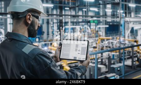 Ingénieur d'usine de voiture dans l'uniforme de travail à l'aide d'un ordinateur tablette avec des plans et des schémas électriques. Spécialiste masculin en usine de fabrication industrielle automobile travaillant sur la production automobile. Banque D'Images