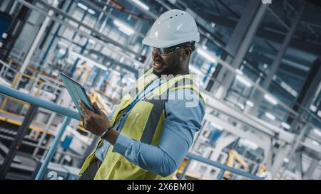 Ingénieur d'usine de voiture afro-américaine dans l'ordinateur de tablette d'utilisation de veste de haute visibilité. Usine de fabrication industrielle automobile produisant un véhicule avec technologie automatisée. Coup d'angle néerlandais Banque D'Images