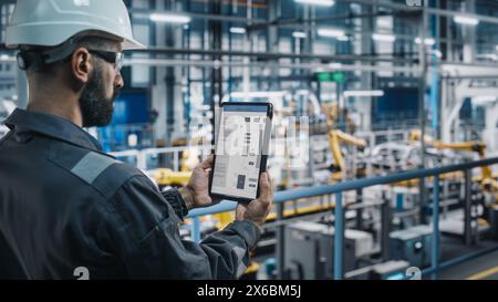 Ingénieur d'usine de voiture dans l'uniforme de travail à l'aide d'un ordinateur tablette avec des plans et des schémas électriques. Spécialiste masculin en usine de fabrication industrielle automobile travaillant sur la production automobile. Banque D'Images