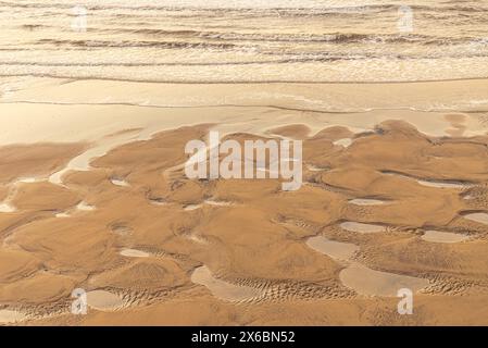 Motifs laissés dans le sable avec des bassins d'eau après une marée nette sur une plage. Les vagues roulent jusqu'au rivage. Banque D'Images