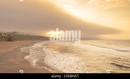 Une scène côtière le soir avec une lumière dorée reflétant de la mer. Les vagues roulent vers le rivage et la lumière du soleil s'échappe d'un trou dans le nuage. Banque D'Images