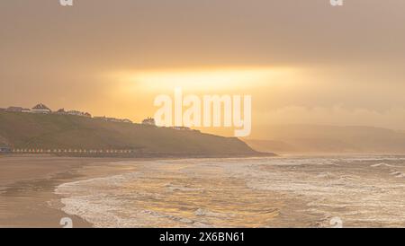 Une scène côtière le soir avec une lumière dorée reflétant de la mer. Les vagues roulent vers le rivage et la lumière du soleil s'échappe d'un trou dans le nuage. Banque D'Images