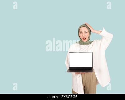 Jeune femme musulmane choquée joyeuse porter le hijab tenir la maquette d'ordinateur portable pc avec l'espace vide de copie d'écran vierge. Tenant sa tête. Banque D'Images